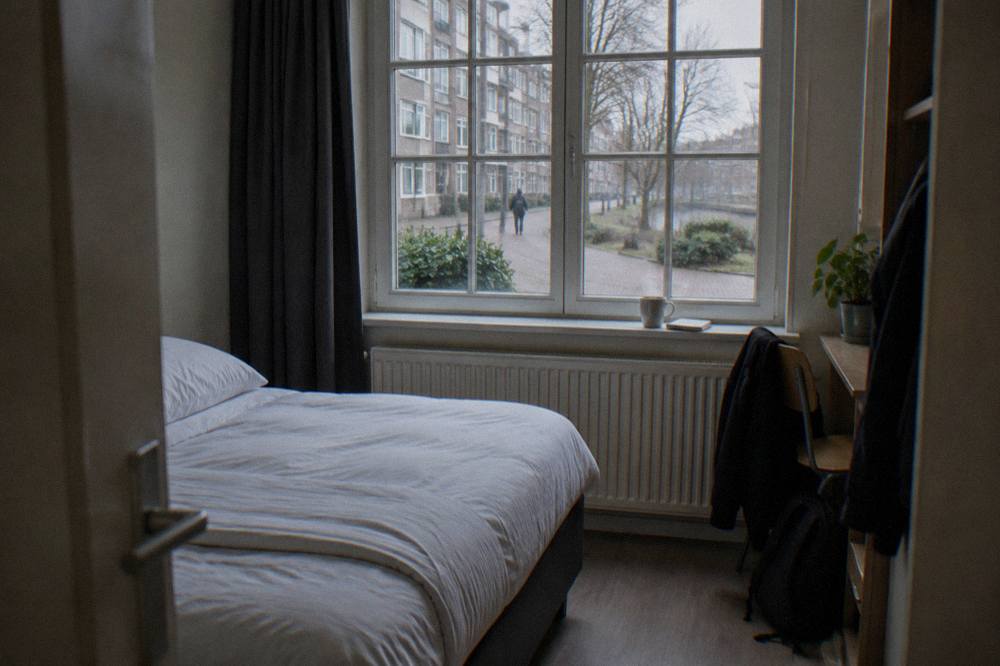A budget hotel room interior with a neatly made bed and a window overlooking an Amsterdam street